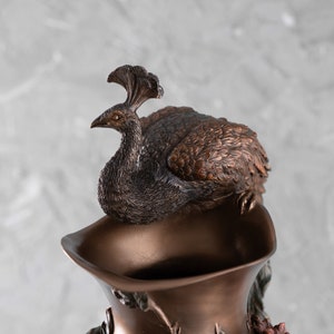 Puede incluir: Un jarr&oacute;n de bronce con un dise&ntilde;o de pavo real. El pavo real est&aacute; posado en la parte superior del jarr&oacute;n y tiene las alas extendidas. El jarr&oacute;n est&aacute; decorado con detalles florales.