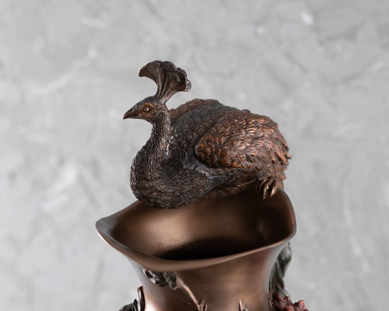 Puede incluir: Un jarr&oacute;n de bronce con un dise&ntilde;o de pavo real. El pavo real est&aacute; posado en la parte superior del jarr&oacute;n y tiene las alas extendidas. El jarr&oacute;n est&aacute; decorado con detalles florales.