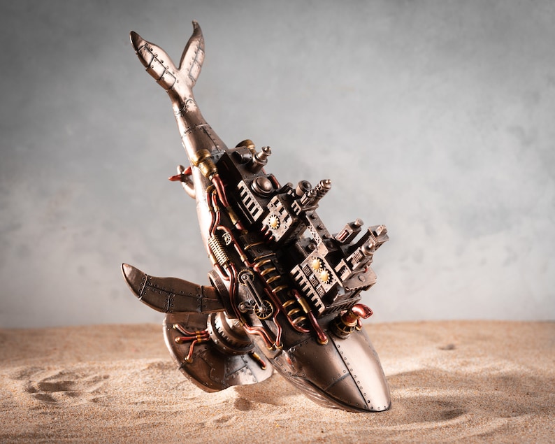 Op de afbeelding: Een steampunk walvisfiguur met ingewikkelde details, gemaakt van metaal en geschilderd in brons- en gouden tinten. De walvis ligt op zijn zij met zijn staart omhoog in de lucht. Het beeldje staat op een zanderige ondergrond.