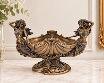 Puede incluir: Un cuenco decorativo de tono bronce con dos figuras de sirenas a cada lado. El cuenco central tiene forma de gran vieira y se apoya sobre una base con detalles fluidos que recuerdan a las olas. Cada sirena lleva un corpiño adornado con conchas.