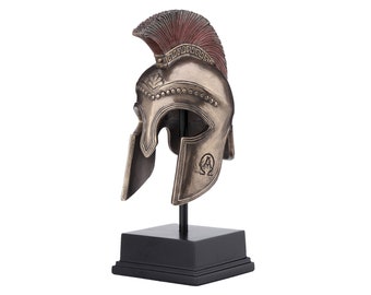 May include: A bronze-colored Greek warrior helmet with a red plume on a black stand. The helmet has a raised design and the Greek letter Omega on the side.