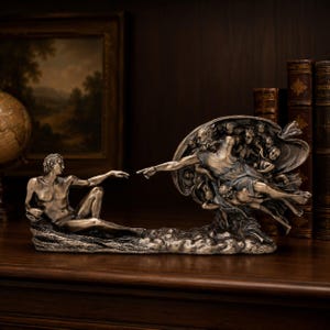 May include: A bronze-toned sculpture depicting the Creation of Adam, featuring a reclining figure reaching toward a floating figure surrounded by cherubs. The detailed metallic finish highlights the muscular forms and flowing drapery of the figures.