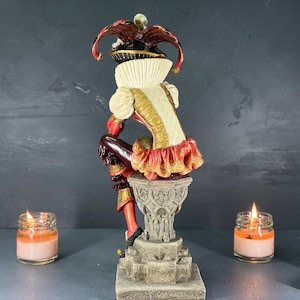 Puede incluir: Figura decorativa de un buf&oacute;n con un disfraz colorido, posada sobre un pedestal de piedra. El buf&oacute;n lleva un sombrero rojo y negro, un cuello con volantes blancos y un atuendo con adornos dorados. La figura se encuentra sobre un fondo oscuro.