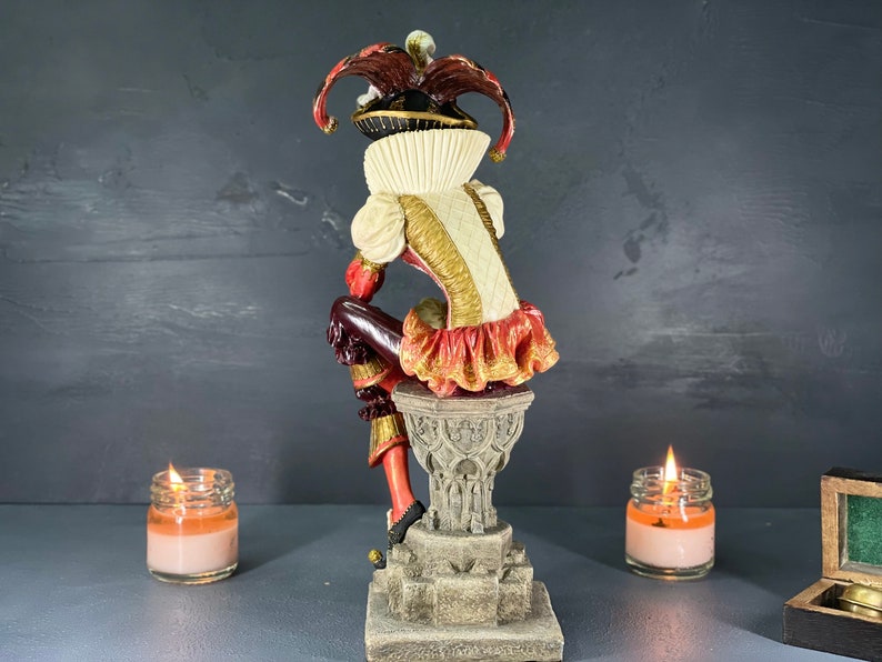 Puede incluir: Figura decorativa de un buf&oacute;n con un disfraz colorido, posada sobre un pedestal de piedra. El buf&oacute;n lleva un sombrero rojo y negro, un cuello con volantes blancos y un atuendo con adornos dorados. La figura se encuentra sobre un fondo oscuro.