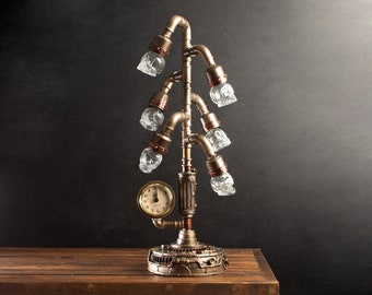 May include: A steampunk-style table lamp with a clock face and six skull-shaped light fixtures. The lamp is made of metal pipes and has a bronze finish.