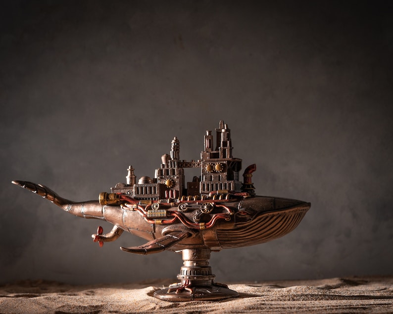 Op de afbeelding: Een steampunk-stijl walvisbeeld met ingewikkelde details en tandwielen. De walvis is gemaakt van metaal en heeft een stad op zijn rug. Het beeld staat op een zanderige ondergrond.
