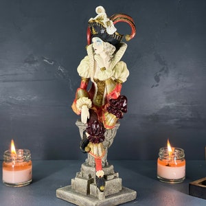 Puede incluir: Figura decorativa de un buf&oacute;n sentado con un disfraz colorido. La figura tiene un sombrero negro con detalles dorados y un atuendo rojo y dorado. Dos velas encendidas en frascos de vidrio flanquean la figura, con una peque&ntilde;a caja de madera.