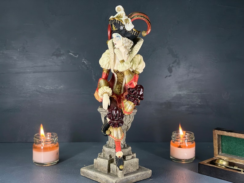 Puede incluir: Figura decorativa de un buf&oacute;n sentado con un disfraz colorido. La figura tiene un sombrero negro con detalles dorados y un atuendo rojo y dorado. Dos velas encendidas en frascos de vidrio flanquean la figura, con una peque&ntilde;a caja de madera.