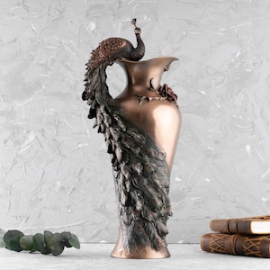 Puede incluir: Un jarr&oacute;n decorativo de bronce con un dise&ntilde;o de pavo real. El jarr&oacute;n es alto y delgado con una boca ancha. El pavo real est&aacute; posado en la parte superior del jarr&oacute;n y sus plumas de la cola est&aacute;n extendidas detr&aacute;s de &eacute;l.