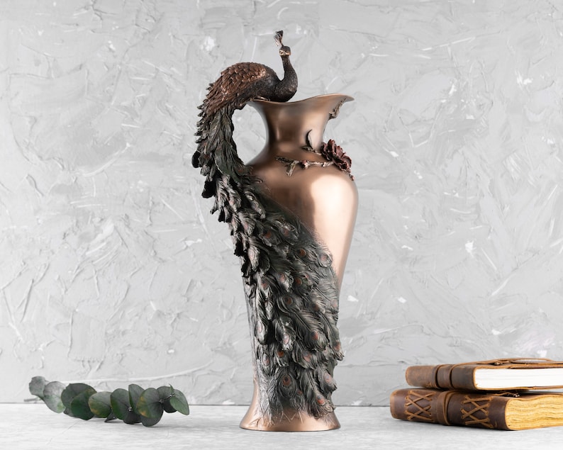 Puede incluir: Un jarr&oacute;n decorativo de bronce con un dise&ntilde;o de pavo real. El jarr&oacute;n es alto y delgado con una boca ancha. El pavo real est&aacute; posado en la parte superior del jarr&oacute;n y sus plumas de la cola est&aacute;n extendidas detr&aacute;s de &eacute;l.