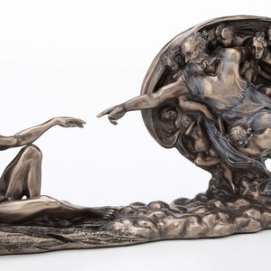 May include: A bronze-toned sculpture depicting the Creation of Adam, featuring a reclining figure reaching toward a bearded figure surrounded by cherubs. The detailed metallic finish highlights the muscular forms and flowing drapery of the figures.