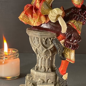 Puede incluir: Figura detallada de una persona con atuendo renacentista, con colores rojo, dorado y burdeos. La figura est&aacute; posada sobre un pedestal decorativo. Una vela encendida en un tarro de cristal est&aacute; en primer plano.