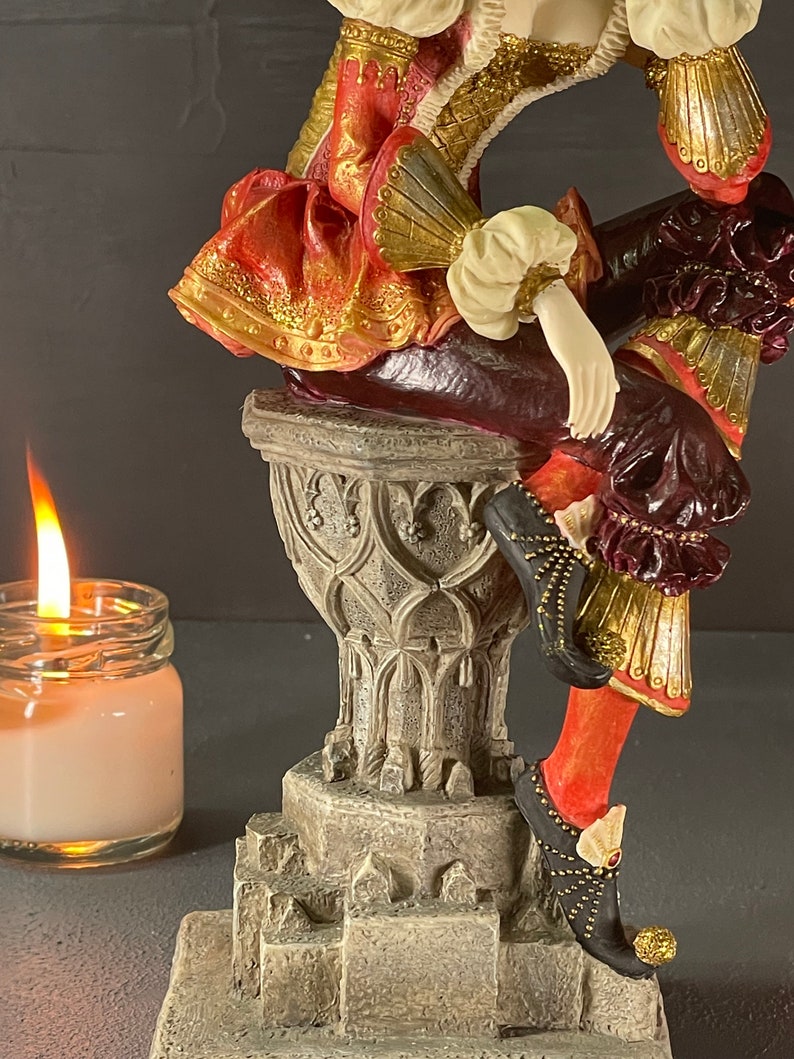 Puede incluir: Figura detallada de una persona con atuendo renacentista, con colores rojo, dorado y burdeos. La figura est&aacute; posada sobre un pedestal decorativo. Una vela encendida en un tarro de cristal est&aacute; en primer plano.