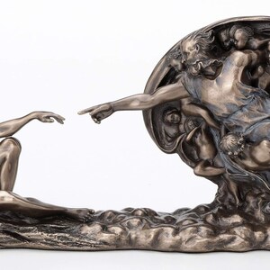 May include: A bronze-toned sculpture depicting the Creation of Adam, featuring a reclining figure reaching out to touch the outstretched finger of a floating figure surrounded by a group of smaller figures within a circular, draped form.