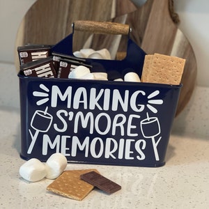 May include: A blue metal caddy with a wooden handle, filled with marshmallows, graham crackers, and chocolate bars. The caddy has the text "Making S'more Memories" printed on it.