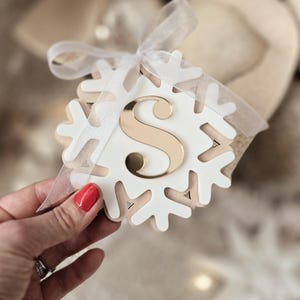 May include: A white snowflake-shaped decoration with a gold letter "S" at its centre, tied with a sheer white ribbon. The decoration is held in a hand with red nail polish. The background is blurred, suggesting a cosy, festive theme.