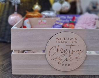 Pode incluir: Uma caixa de madeira com uma placa redonda de madeira que diz "Willow & Felicity's Christmas Eve Box". A caixa está cheia de presentes e está sentada em frente a uma árvore de Natal.
