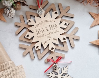May include: A wooden snowflake ornament with the text "Our First Christmas" and the names "Jessi & Fred" and the text "Together at 43 Main Street 2021". The ornament is hanging from a red ribbon and has a small bell attached to the bottom.