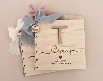 May include: A wooden book with a light wood finish. The cover has the letter "T" and the name "Thomas" written in cursive. The text "On your first birthday" is also written on the cover. The book is decorated with blue and pink ribbons.