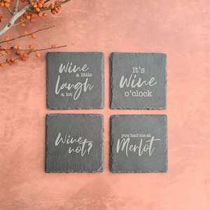 May include: Four square slate coasters with white lettering. The coasters have the following phrases: "Wine a little, laugh a lot", "It's wine o'clock", "Wine not?", and "You had me at Merlot". The coasters are arranged on a pink surface.