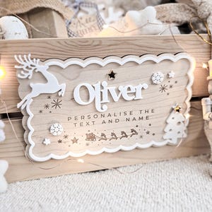 May include: Wooden sign with the name "Oliver" in white, featuring a reindeer, snowflakes, and a Christmas tree. The sign reads "Personalise the text and name" with a Santa and reindeer sleigh design. Decorative lights and other Christmas decorations are in the background.