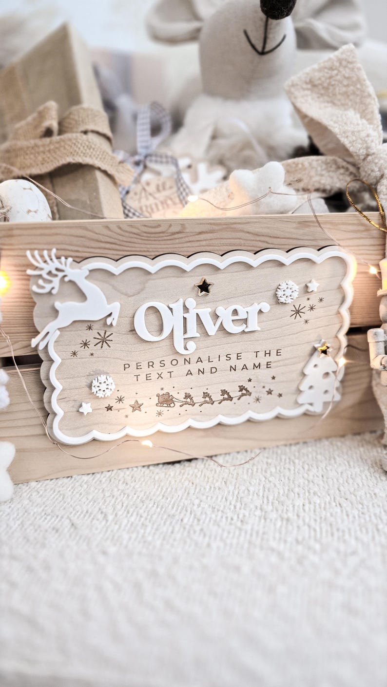 May include: Wooden sign with the name "Oliver" in white, featuring a reindeer, snowflakes, and a Christmas tree. The sign reads "Personalise the text and name" with a Santa and reindeer sleigh design. Decorative lights and other Christmas decorations are in the background.