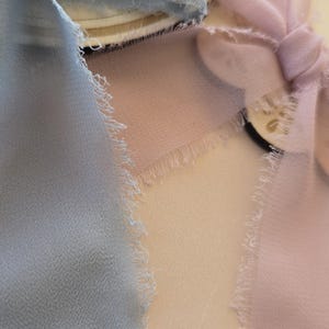 Może przedstawiać: Zbliżenie na trzy pr&oacute;bki tkanin w pastelowych kolorach: jasnoniebieskim, bladym r&oacute;żu i kremowym beżu. Każda tkanina ma postrzępiony brzeg, dodający delikatnej faktury. Miękki, zwiewny materiał to prawdopodobnie jedwab lub podobny lekki materiał.