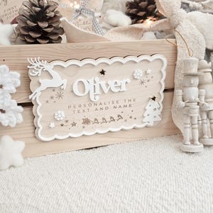 May include: A wooden sign with the name "Oliver" in white, surrounded by decorative elements like a reindeer, snowflakes, stars, and a Christmas tree. The sign includes the text "Personalise the text and name". The sign is light brown with a scalloped edge.