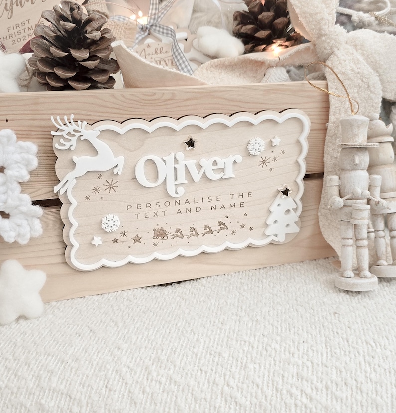 May include: A wooden sign with the name "Oliver" in white, surrounded by decorative elements like a reindeer, snowflakes, stars, and a Christmas tree. The sign includes the text "Personalise the text and name". The sign is light brown with a scalloped edge.