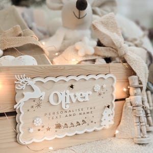May include: A wooden sign with the name "Oliver" in white, surrounded by decorative elements like a reindeer, snowflakes, and a Christmas tree. The sign is part of a Christmas-themed display.