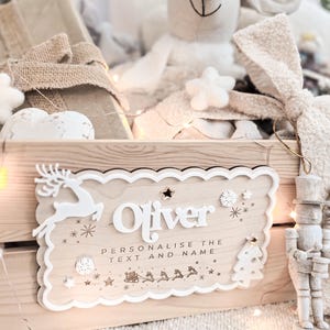 May include: A wooden crate with a personalized sign that reads "Oliver". The sign has a white border with decorative elements like a reindeer, snowflakes, and a Christmas tree. The crate is filled with gifts and a plush mouse.