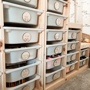 May include: A wooden toy storage unit with multiple drawers labelled with wooden circles. The labels include "Books", "Colours", "Puzzles", "Music", and "3+1=2".