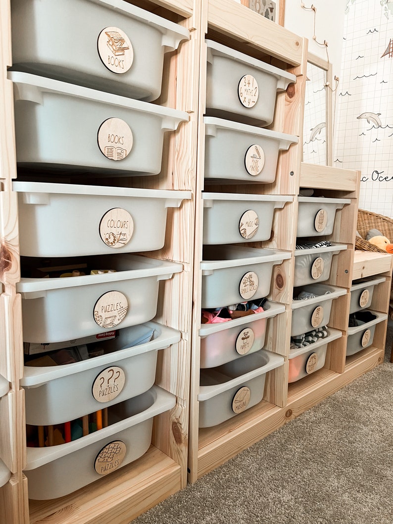 May include: A wooden toy storage unit with multiple drawers labelled with wooden circles. The labels include "Books", "Colours", "Puzzles", "Music", and "3+1=2".