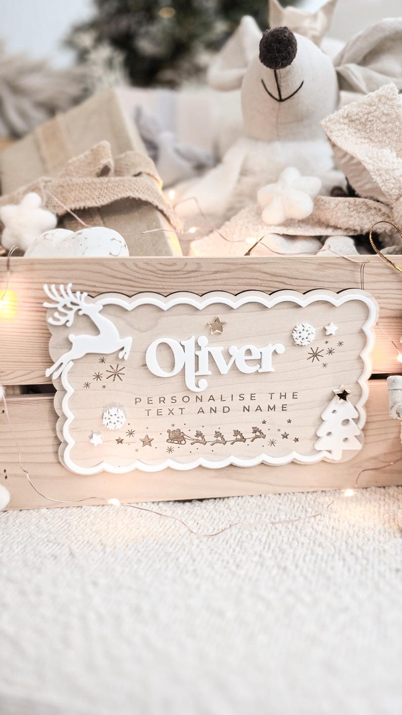 May include: Wooden sign with the name "Oliver" in white, featuring a reindeer, stars, and a Christmas tree design. The sign includes the text "Personalise the text and name". The sign is on a wooden surface.