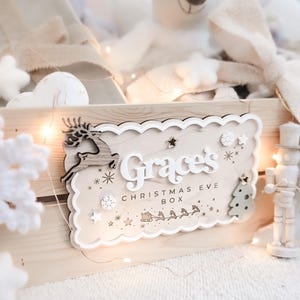 May include: A wooden Christmas Eve box with a decorative sign that reads "Grace's CHRISTMAS EVE BOX". The sign features a reindeer, stars, and a Christmas tree. The box is filled with soft toys and decorations.