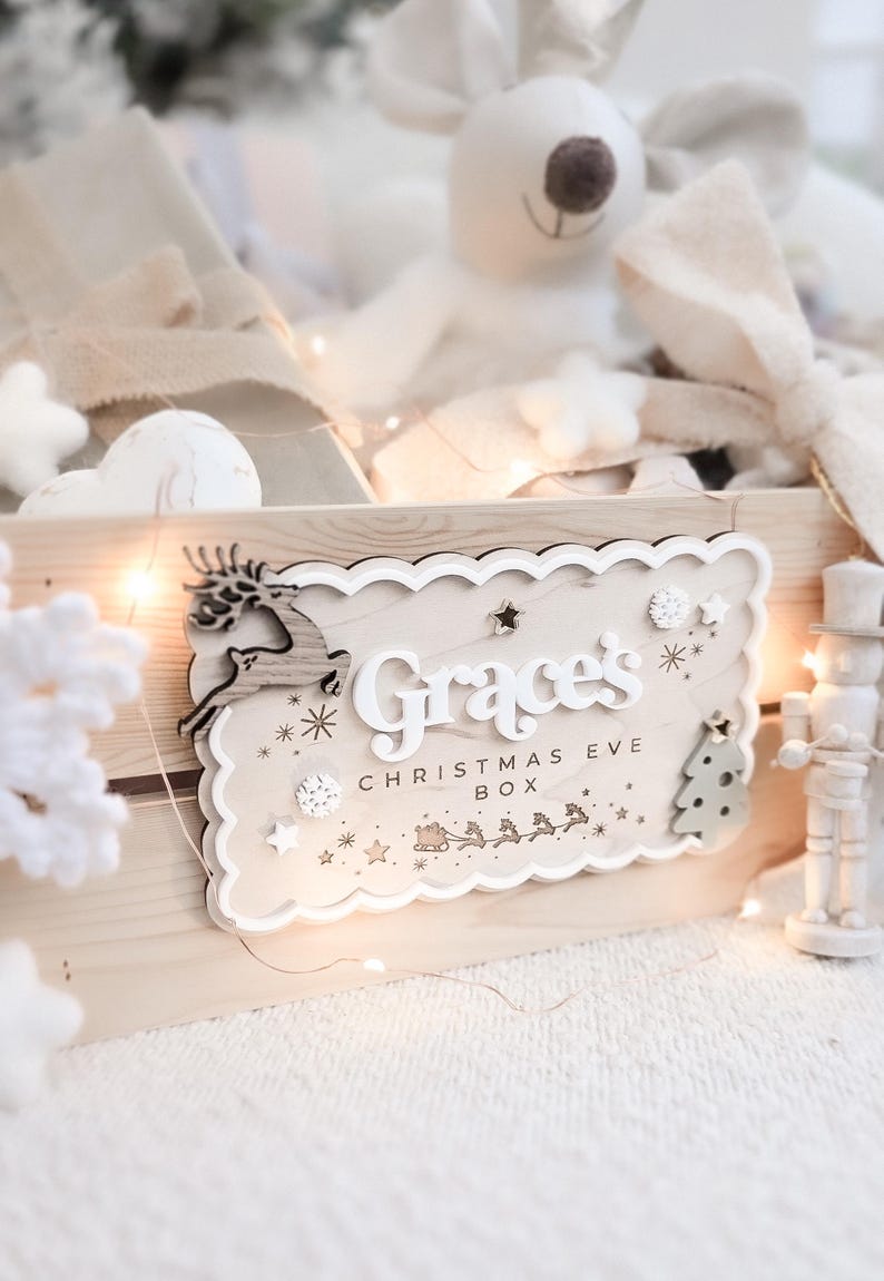 May include: A wooden Christmas Eve box with a decorative sign that reads "Grace's CHRISTMAS EVE BOX". The sign features a reindeer, stars, and a Christmas tree. The box is filled with soft toys and decorations.