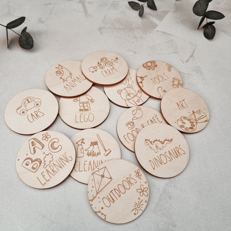 May include: A set of 12 wooden circle tags with various illustrations and text. The tags include images of cars, dinosaurs, a farm, tools, a cafe, art supplies, a house, a spray bottle, a kite, a toy bear, letters, and an animal. The text on the tags includes "Cars", "Dinosaurs", "Farm", "Tools", "Food & Cafe", "Art", "Lego", "Cleaning", "Outdoors", "Toys", "A B C Learning", and "Animal".