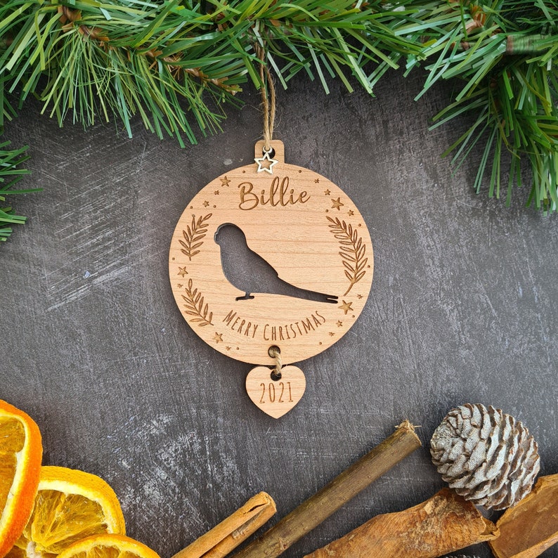 May include: A wooden Christmas ornament with a bird silhouette, the name "Billie" and the text "Merry Christmas". A smaller heart-shaped ornament with the year "2021" hangs below.