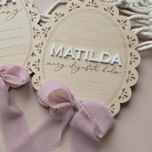 Może przedstawiać: Drewniany znak z falistą krawędzią z napisem "MATILDA" w białych, wypukłych literach. Poniżej kursywą napisano imię "Mary Elizabeth Helm". U dołu zawiązana jest jasno-r&oacute;żowa wstążka.