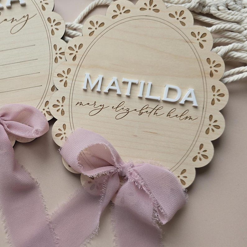 Może przedstawiać: Drewniany znak z falistą krawędzią z napisem "MATILDA" w białych, wypukłych literach. Poniżej kursywą napisano imię "Mary Elizabeth Helm". U dołu zawiązana jest jasno-r&oacute;żowa wstążka.