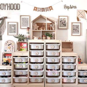 May include: A white wooden storage unit with 16 clear plastic drawers. Each drawer has a round wooden label with a different word on it. The unit is in a room with a white wall and a wooden dollhouse. The words on the labels include "Boyhood", "Explore", and "Arlin".