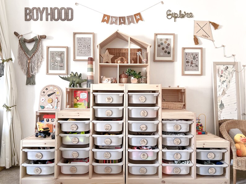 May include: A white wooden storage unit with 16 clear plastic drawers. Each drawer has a round wooden label with a different word on it. The unit is in a room with a white wall and a wooden dollhouse. The words on the labels include "Boyhood", "Explore", and "Arlin".