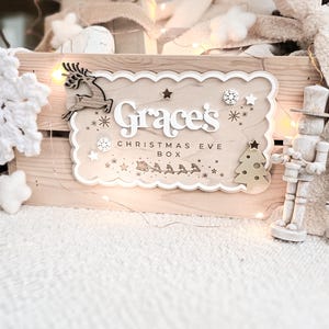 May include: A wooden Christmas Eve box with the name "Grace's" and the text "Christmas Eve Box" in white. The box is decorated with a reindeer, stars, a Christmas tree, and a sleigh with reindeer. A wooden toy soldier stands to the right.