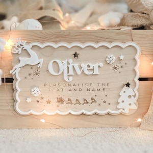 May include: Wooden sign with the name "Oliver" in white, featuring a reindeer, snowflakes, stars, and a Christmas tree. The sign includes the text "Personalise the text and name" and a sleigh with reindeer.