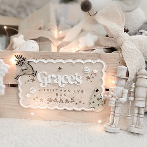 May include: A wooden Christmas Eve box with the name "Grace's" and the text "Christmas Eve Box" in white lettering. The box is decorated with a reindeer, stars, snowflakes, and a Christmas tree. A plush mouse and nutcrackers are also visible.