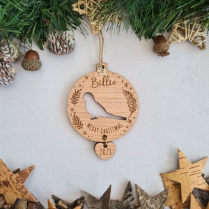 May include: A wooden Christmas ornament shaped like a circle with a bird silhouette, the words "Billie Merry Christmas" and a star design. A smaller heart-shaped ornament with the year "2021" hangs from the bottom.
