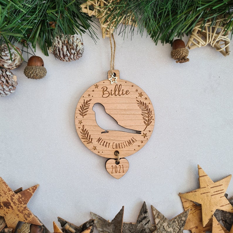 May include: A wooden Christmas ornament shaped like a circle with a bird silhouette, the words "Billie Merry Christmas" and a star design. A smaller heart-shaped ornament with the year "2021" hangs from the bottom.