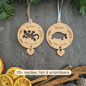 May include: Two wooden Christmas decorations featuring a cut-out gecko and a turtle. The decorations are personalised with the names "Gizmo" and "Trevor" and the year "2021".