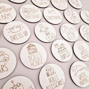 May include: A set of 16 wooden circle toy labels with various images and text, including "Farm", "Dress Up", "Dinosaurs", "Threading", "Lego", "Dolls House", "Duplo", "Water Animals", and "Dolls Accessories".