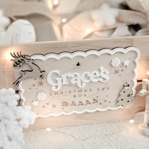 May include: A wooden Christmas Eve box with a scalloped edge, featuring the name "Grace's" in white letters. The box is decorated with a reindeer, stars, snowflakes, and a Christmas tree. The words "CHRISTMAS EVE BOX" are also visible.